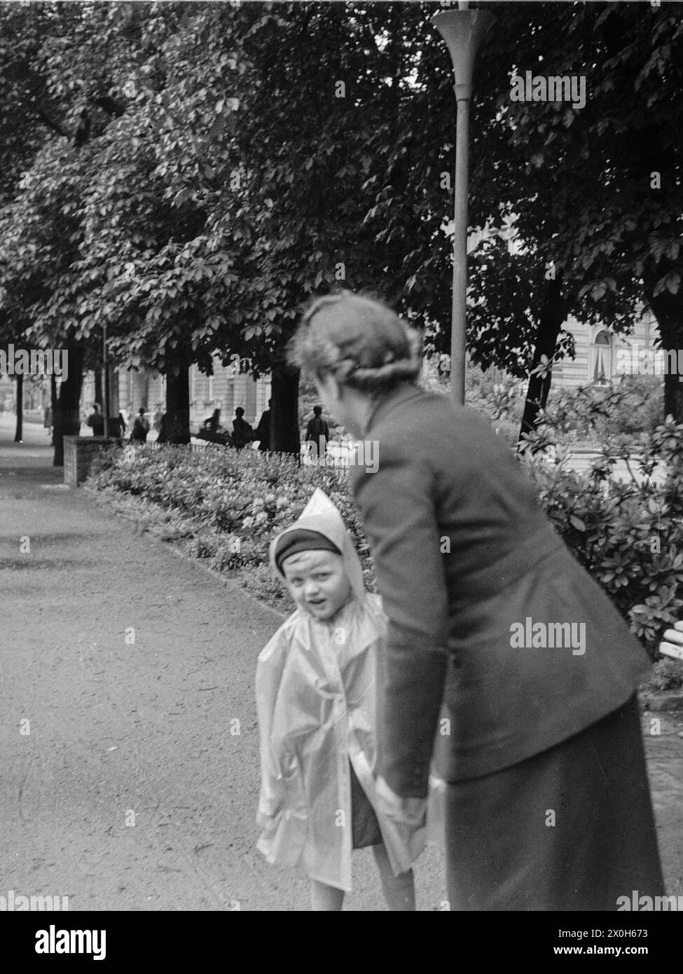 Une mère emmène son jeune fils se promener dans un parc en ville. L'enfant porte une cape de pluie. [traduction automatique] Banque D'Images