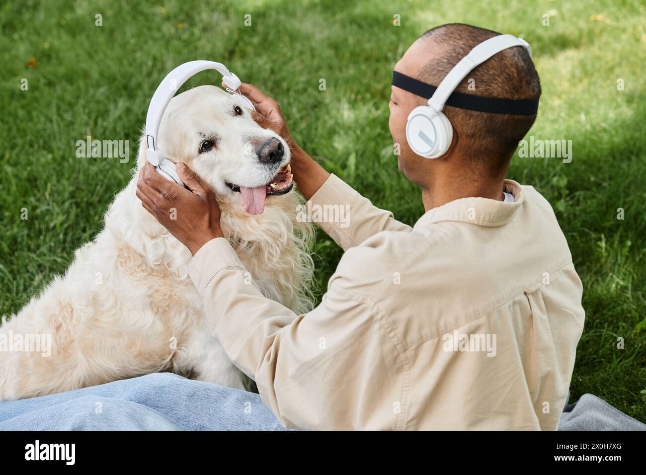 Un homme diversifié atteint du syndrome de la myasthénie grave est assis sur l'herbe, caressant joyeusement son fidèle chien Labrador tandis que les deux portent des écouteurs. Banque D'Images