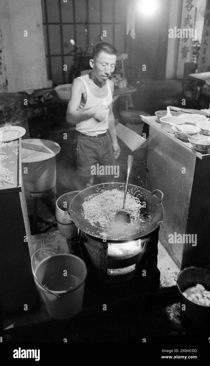 Un homme se tient dans une poêle à wok à Singapour. La cuisine est aménagée à l'extérieur par le bord de la route. Une lampe donne de la lumière dans l'obscurité [traduction automatique] Banque D'Images