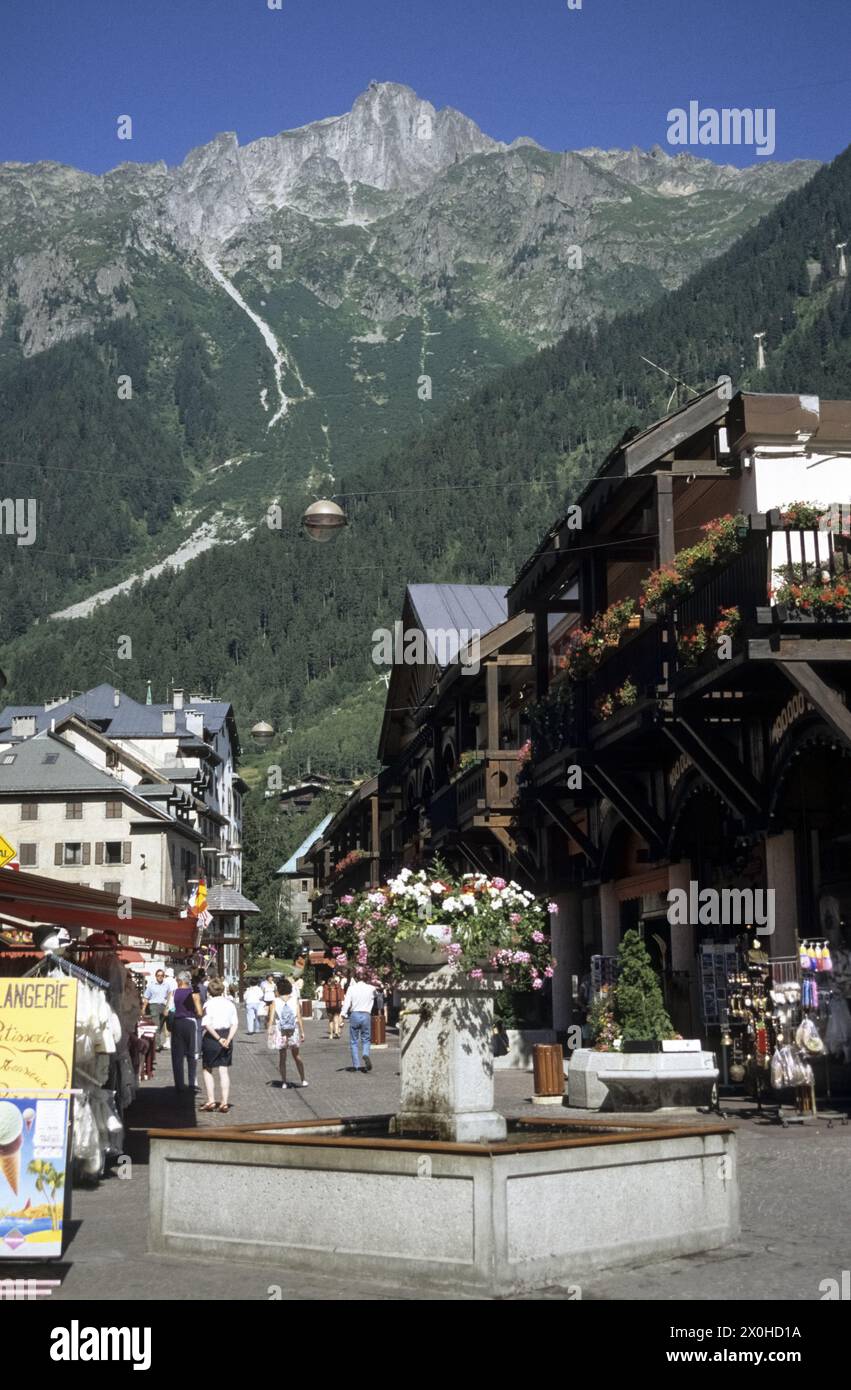 La zone piétonne avec une fontaine décorée de fleurs, de magasins et de quelques passants par une chaude journée d'été. Le BrÃ©vent en arrière-plan. [traduction automatique] Banque D'Images