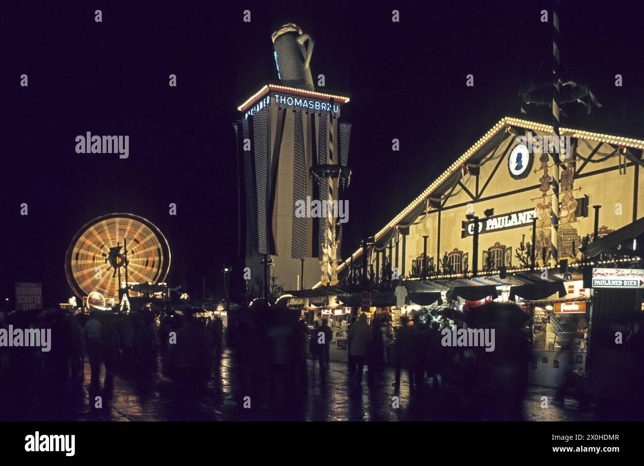 Les gens sur les stands devant la tente du festival Paulaner et une grande roue illuminée la nuit en arrière-plan à l'Oktoberfest de Munich. [traduction automatique] Banque D'Images