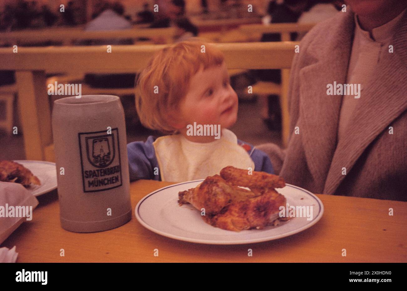 Un petit enfant avec un bavoir est assis dans la tente à bière devant un mug à bière et une assiette avec un demi-poulet pendant l'Oktoberfest à Munich. [traduction automatique] Banque D'Images