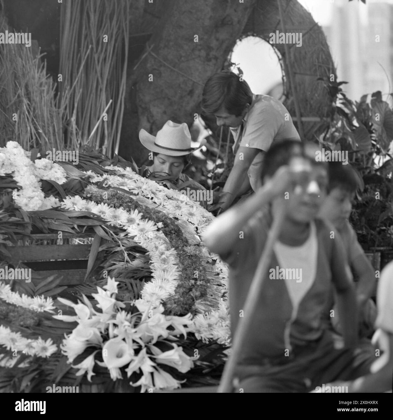 Deux hommes travaillent sur une grande couronne de fleurs à Guernavaca. L'un d'eux porte un chapeau avec un large bord. En arrière-plan vous pouvez voir d'autres matériaux et couronnes finies de fleurs. [traduction automatique] Banque D'Images