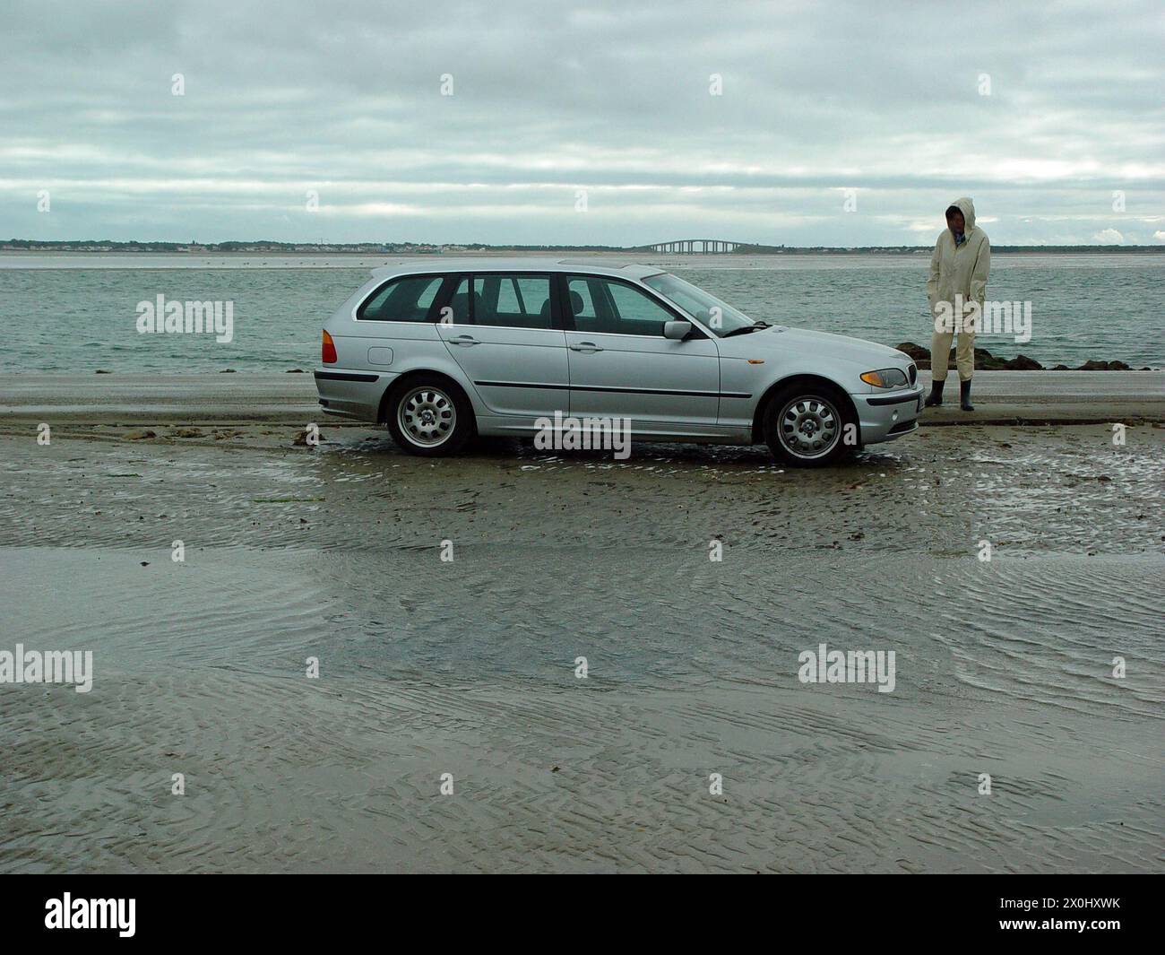 'Vue sur le passage du Gois. Au premier plan se trouve une personne debout à côté d'une BMW au bord de la route. La route est entourée d'eau et de vasières. La personne porte des bottes en caoutchouc et une veste de pluie. En arrière-plan, vous pouvez voir le pont ''Pont du Noirmoutier''. [traduction automatique]' Banque D'Images