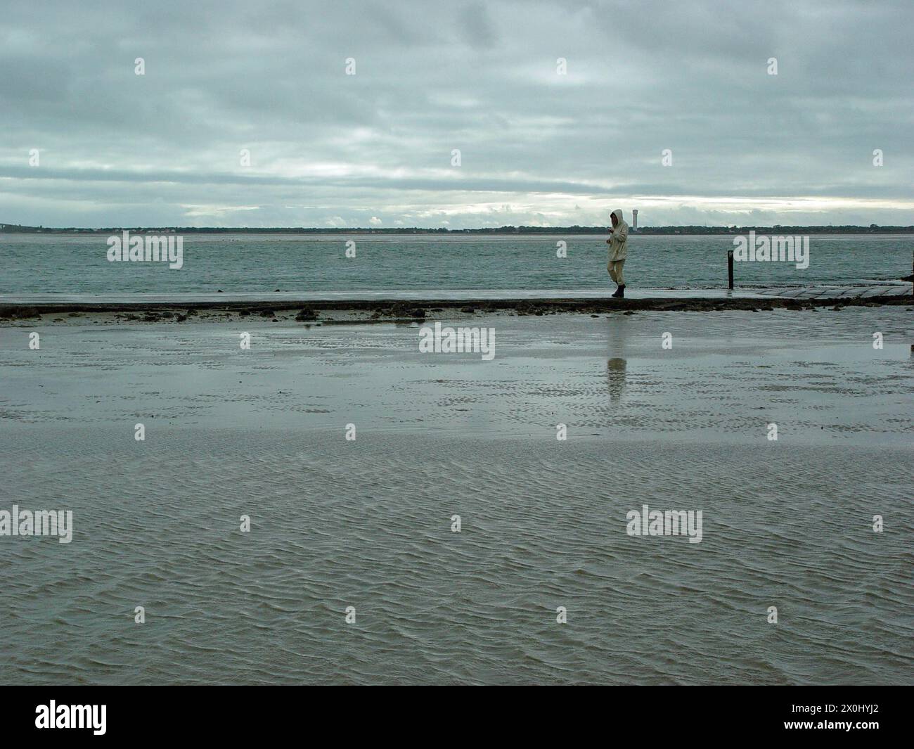 Vue sur le passage du Gois en France. Sur la route presque inondée remblai une femme dans des bottes en caoutchouc et la veste de pluie est sur le chemin. La route est entourée d'eau et de vasières. En arrière-plan vous pouvez voir la côte. [traduction automatique] Banque D'Images