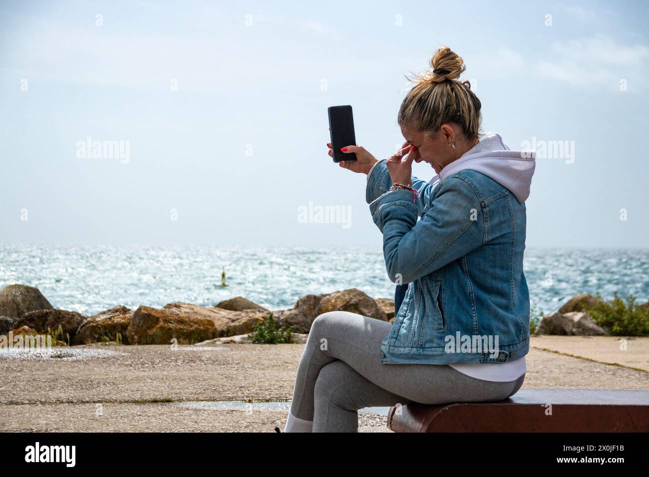 Femme blonde avec chignon en utilisant son téléphone portable tout en profitant de la mer assise sur un banc sur la promenade Banque D'Images