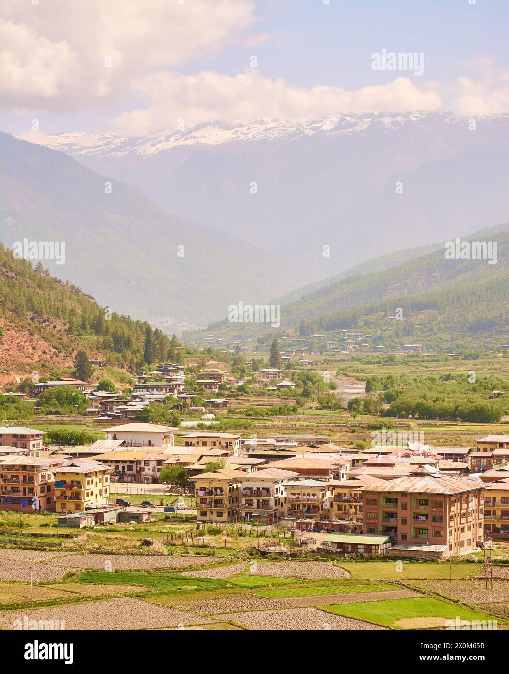 Une vue sur les champs, le logement et Paro Chhu (rivière) dans la ville de Paro dans la vallée de Paro avec des montagnes avec de la neige au sommet au printemps, l'ouest du Bhoutan. Banque D'Images