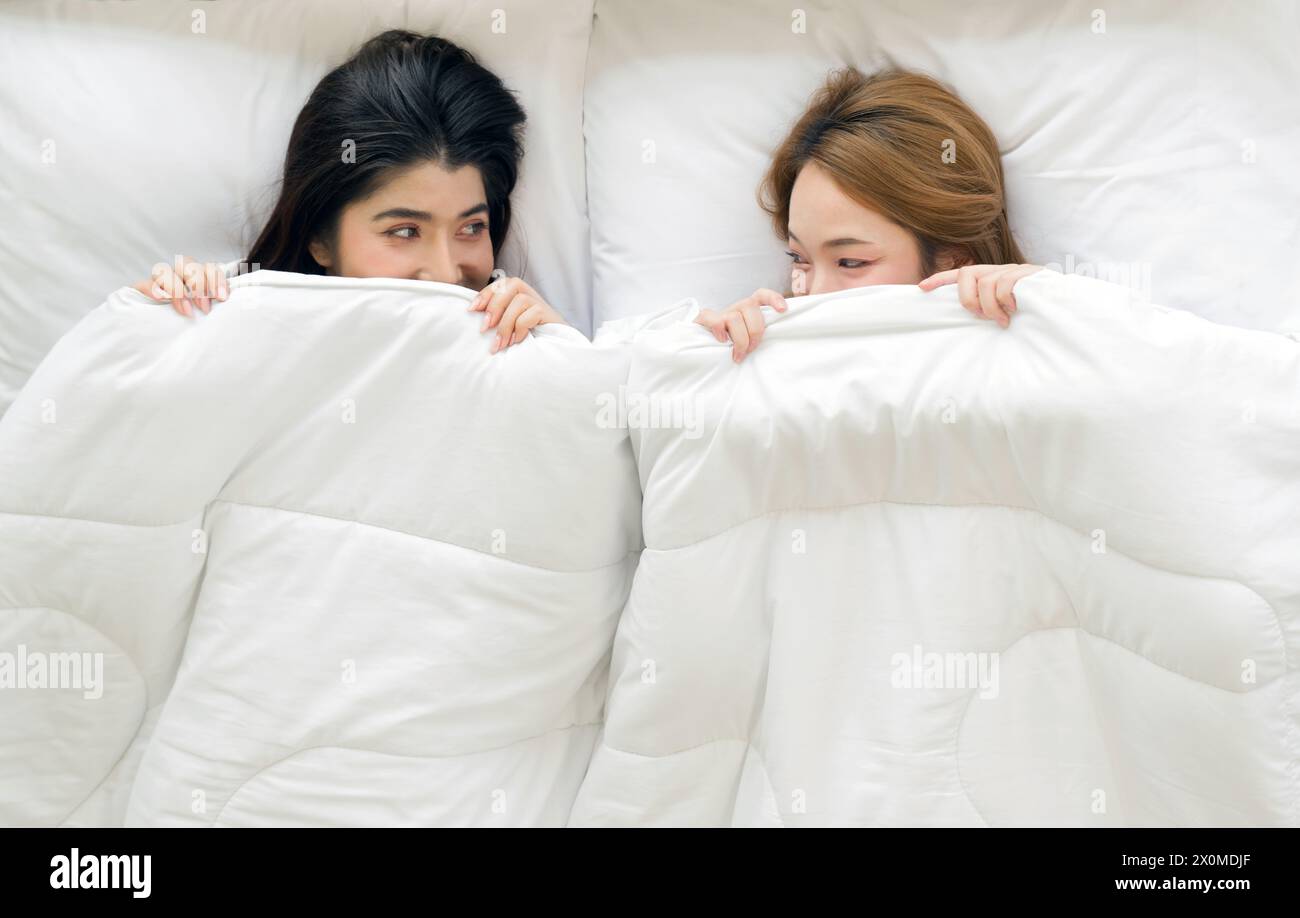 Jeune couple asiatique regarde sous une couverture blanche dans un lit, se regardant avec affection. Banque D'Images