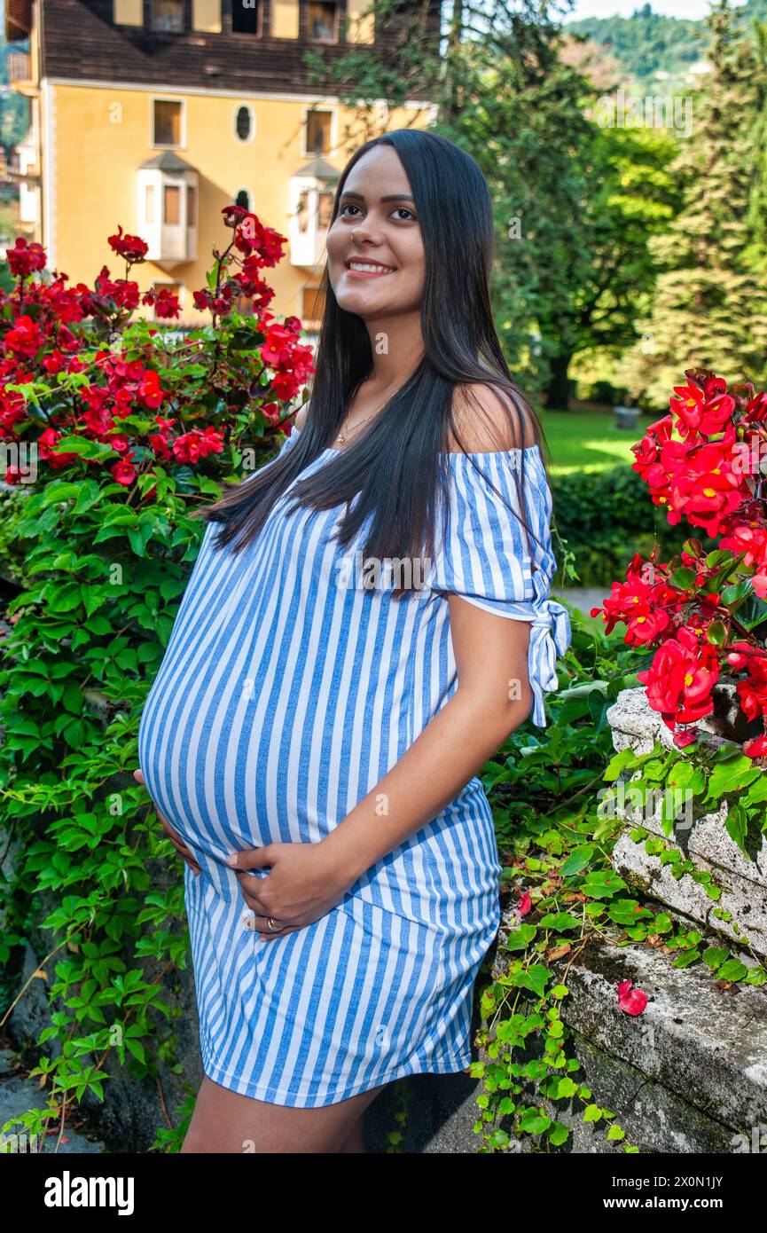 Souriante belle fille sud-américaine enceinte, au milieu des fleurs, portant une longue chemise rayée Banque D'Images
