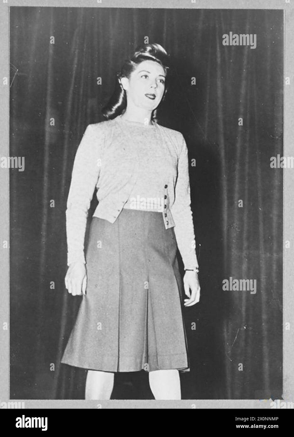 Un mannequin modélise un ensemble jumeau en laine grise avec une jupe plissée écarlate lors d'un salon de robe utilitaire de dépôt de réparation d'avions en 1945. Banque D'Images
