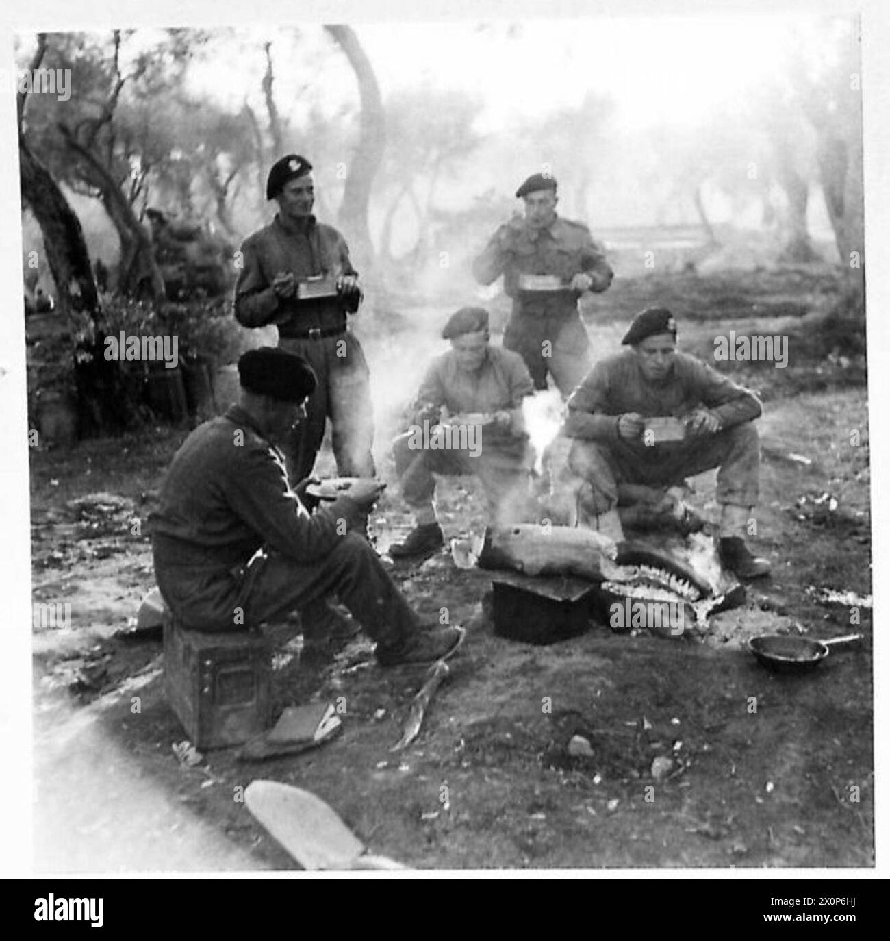 Négatif photographique montrant un officier et l'équipage d'un char Sherman prenant un repas du soir autour d'un feu de bois avec le soleil couchant en arrière-plan. Banque D'Images