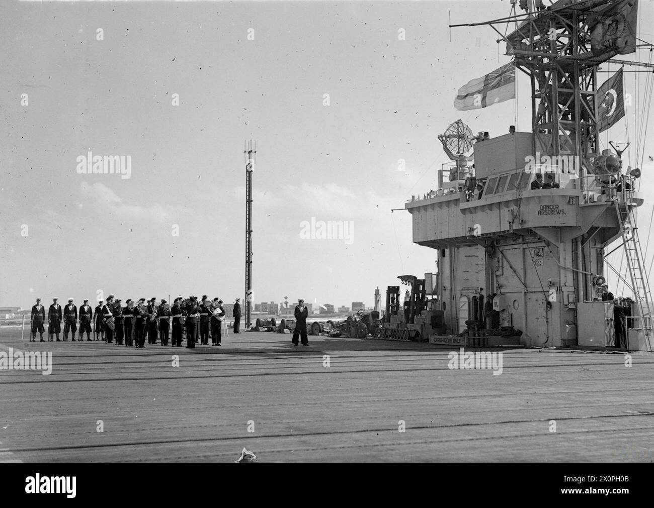 LE ROI FAROUK VISITE LE PORTE-AVIONS D'ESCORTE BRITANNIQUE. MARS 1945, À BORD DU HMS HUNTER, ALEXANDRIE, ÉGYPTE. - L'étalon royal égyptien et l'enseigne volant sur le générique du CHASSEUR avec l'enseigne blanche en tant que roi Farouk, sur le pont, est joué dans le port par le Royal Marine Band Banque D'Images