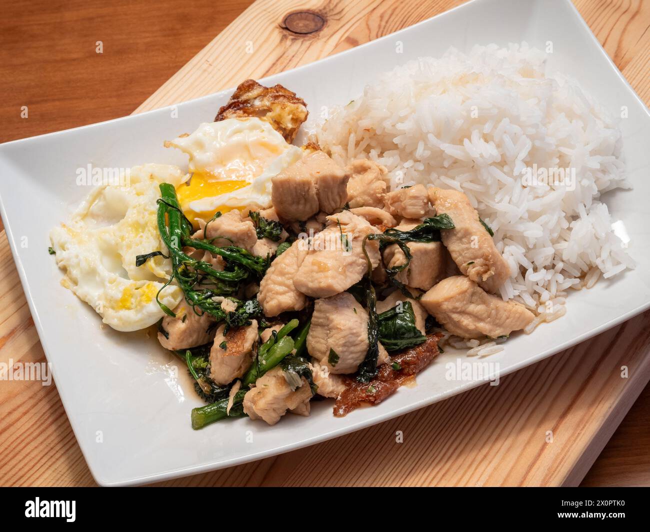 Laissez-vous tenter par les saveurs du poulet au basilic thaïlandais servi avec un œuf parfaitement frit, du broccolini et du riz au jasmin parfumé, élégamment présenté sur un blanc Banque D'Images