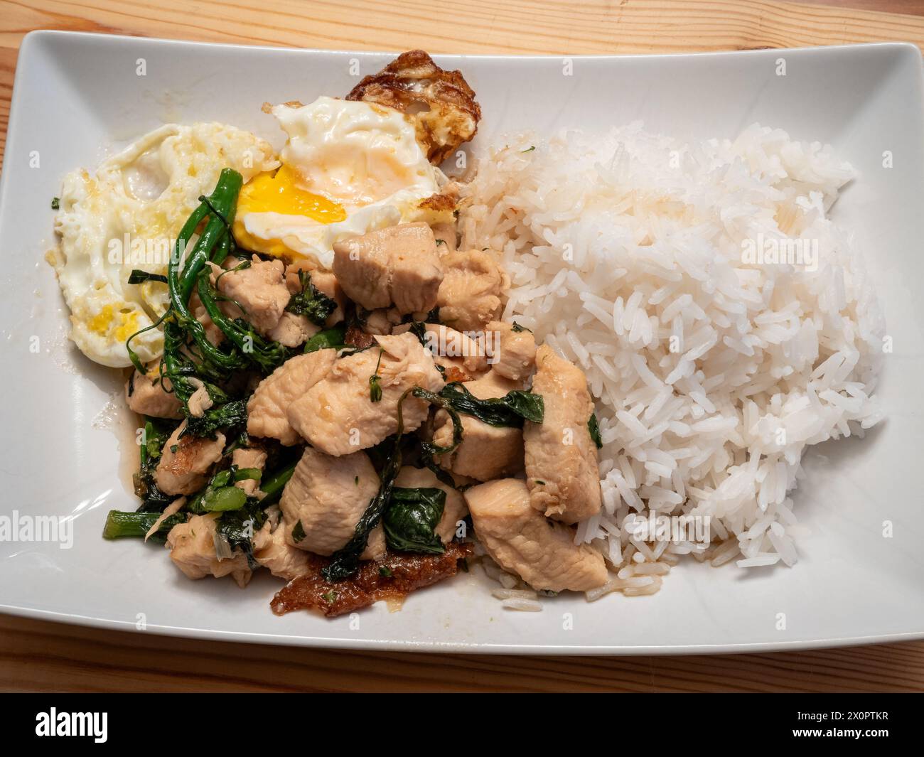 Laissez-vous tenter par les saveurs du poulet au basilic thaïlandais servi avec un œuf parfaitement frit, du broccolini et du riz au jasmin parfumé, élégamment présenté sur un blanc Banque D'Images