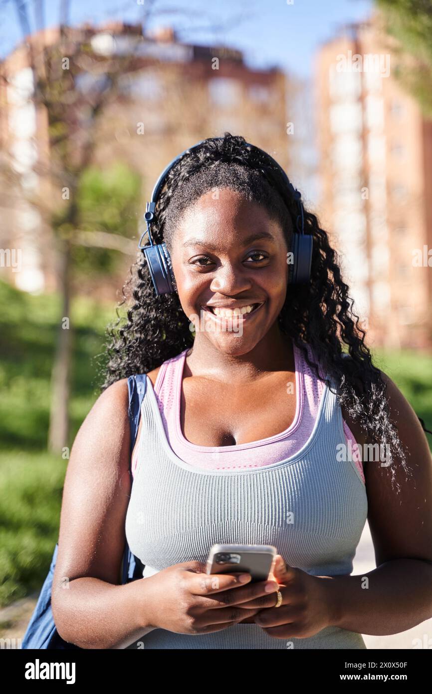 Portrait vertical d'une jeune femme afro-américaine écoutant de la musique avec des écouteurs tout en tenant un smartphone et en regardant l'appareil photo. Banque D'Images