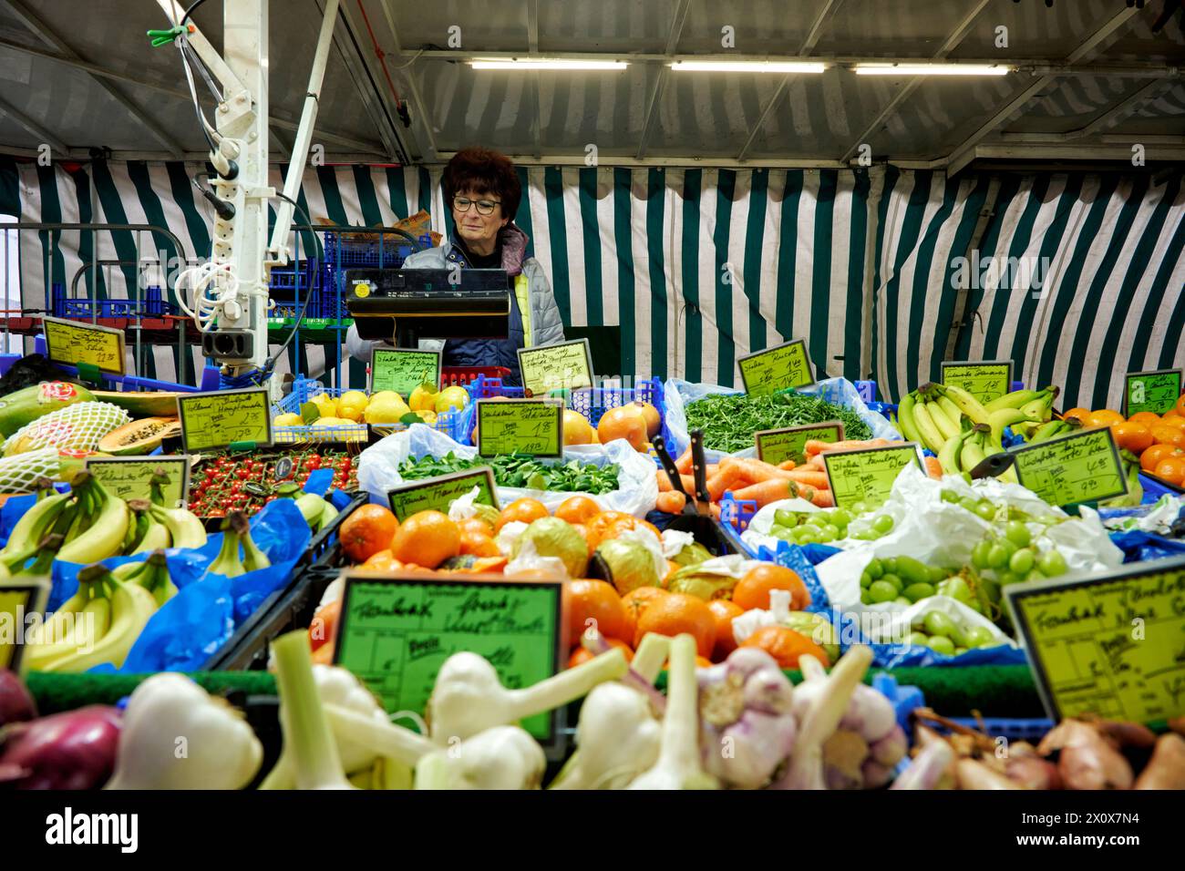 Reichhaltige Obst- und Gemüseauswahl an einem Marktstand. Eine Verkäuferin steht im Hintergrund an einer Waage und wiegt aus. Banque D'Images