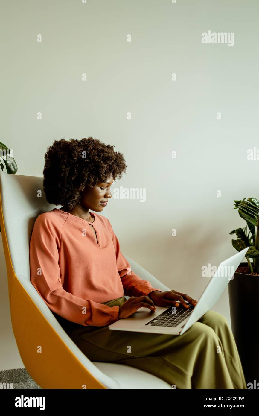 Femme d'affaires afro-américaine animée souriante tout en travaillant sur son ordinateur portable, confortablement assise dans un salon de bureau animé. Banque D'Images