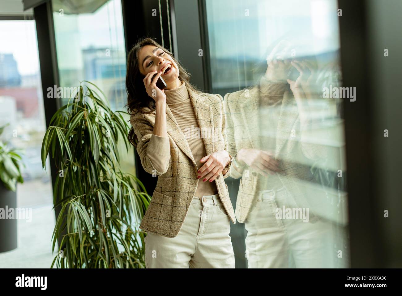 Femme contemplative utilisant un téléphone portable et regarde par une fenêtre, avec une expression réfléchie et des vues sur la ville derrière elle Banque D'Images