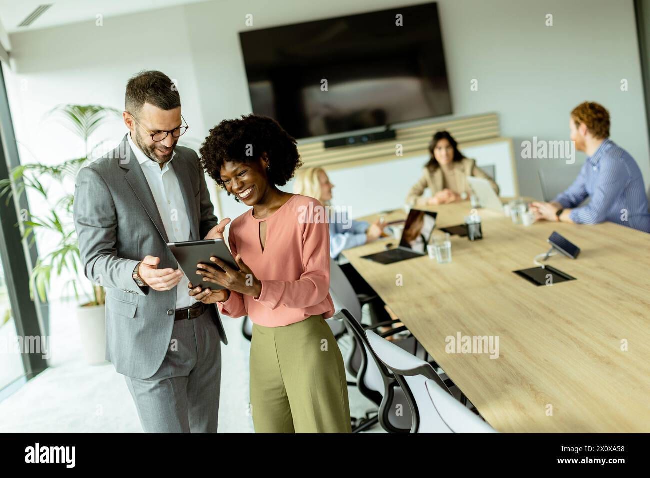 Homme souriant et femme d'affaires afro-américaine appréciant le contenu sur une tablette avec des collègues en arrière-plan Banque D'Images