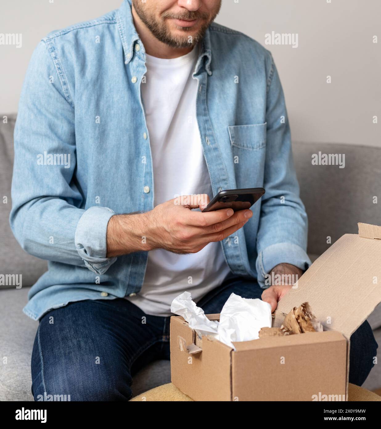 Homme dans une chemise en denim déballe un paquet et prend une photo de son achat, commande internet. Personne partageant des commentaires avec la boutique en ligne en utilisant le sien Banque D'Images