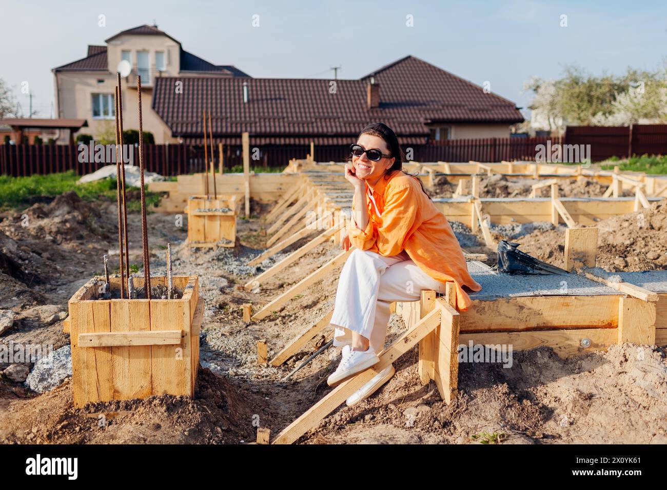 Heureuse femme propriétaire assise sur la fondation en béton pour la nouvelle maison entourée de terrasse en bois en attente de la future maison à construire. Banque D'Images