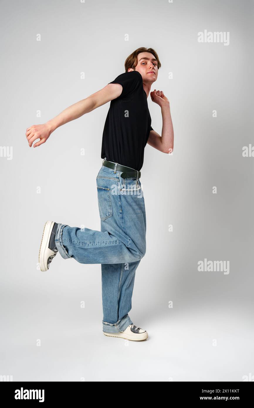 Jeune homme exécutant un mouvement de danse décontracté en jeans et baskets sur un fond gris Banque D'Images