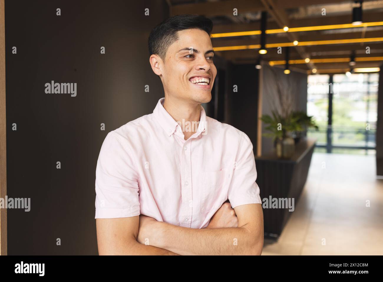 Un jeune homme biracial portant une chemise rose est debout, les bras croisés, riant Banque D'Images