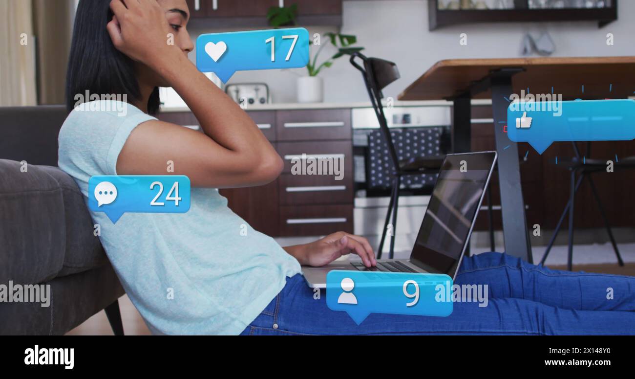 Image des icônes de médias sociaux et des numéros sur la femme biraciale à l'aide d'un ordinateur portable. Concept mondial de médias sociaux et de traitement des données image générée numériquement. Banque D'Images