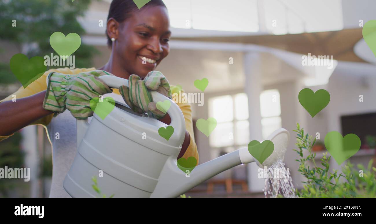 Image de coeurs verts sur la femme afro-américaine heureuse arrosant des plantes Banque D'Images
