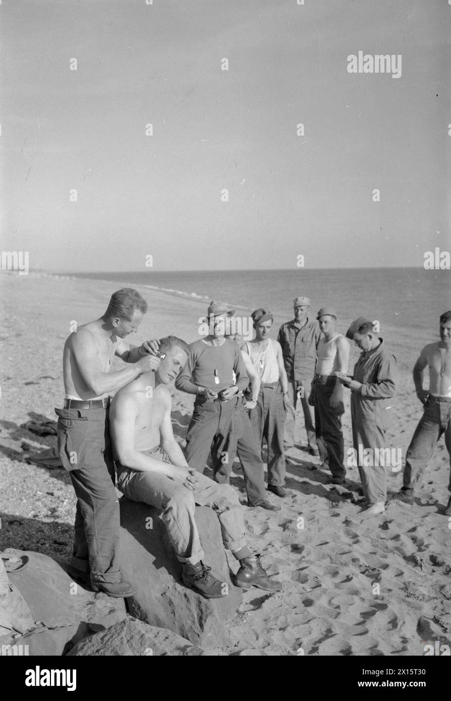 TROUPES AMÉRICAINES DANS UN VILLAGE ANGLAIS : VIE QUOTIDIENNE AVEC LES AMÉRICAINS À BURTON BRADSTOCK, DORSET, ANGLETERRE, Royaume-Uni, 1944 - sur une plage près de leur camp à Burton Bradstock (peut-être Burton Beach ou Burton Freshwater), les troupes américaines visitent le coiffeur au soleil en début de soirée. Barbier amateur T/5 John G Artie (d'Edison Avenue, Menlo Park, New Jersey) coupe les cheveux du T/5 John B Rada (du 103 Stone Avenue, Greenville, South Indiana), qui est assis sur un rocher, à la place d'un fauteuil de barbier. Attendent leur tour : T/4 George Keister (de Fort Wayne, Indiana), T/5 H R Sullivan (de Hodges, Alabama), PFC He Banque D'Images