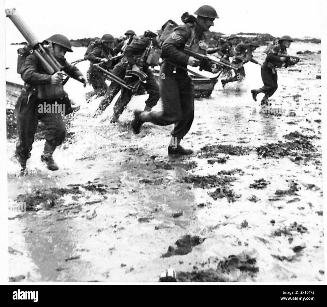 Les hommes armés de pistolets Tommy et de mitrailleuses pratiquent les débarquements sur la plage, se déplaçant à travers la mousse d'eau pour gagner des positions pendant les exercices de l'armée. Banque D'Images