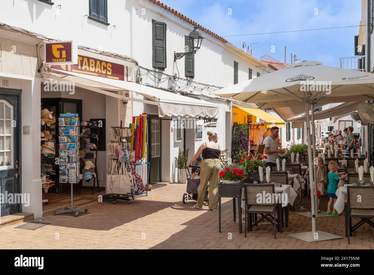 Les touristes marchent dans une rue étroite et confortable avec des restaurants et des boutiques dans le village de Fornells sur l'île espagnole de Minorque. Banque D'Images