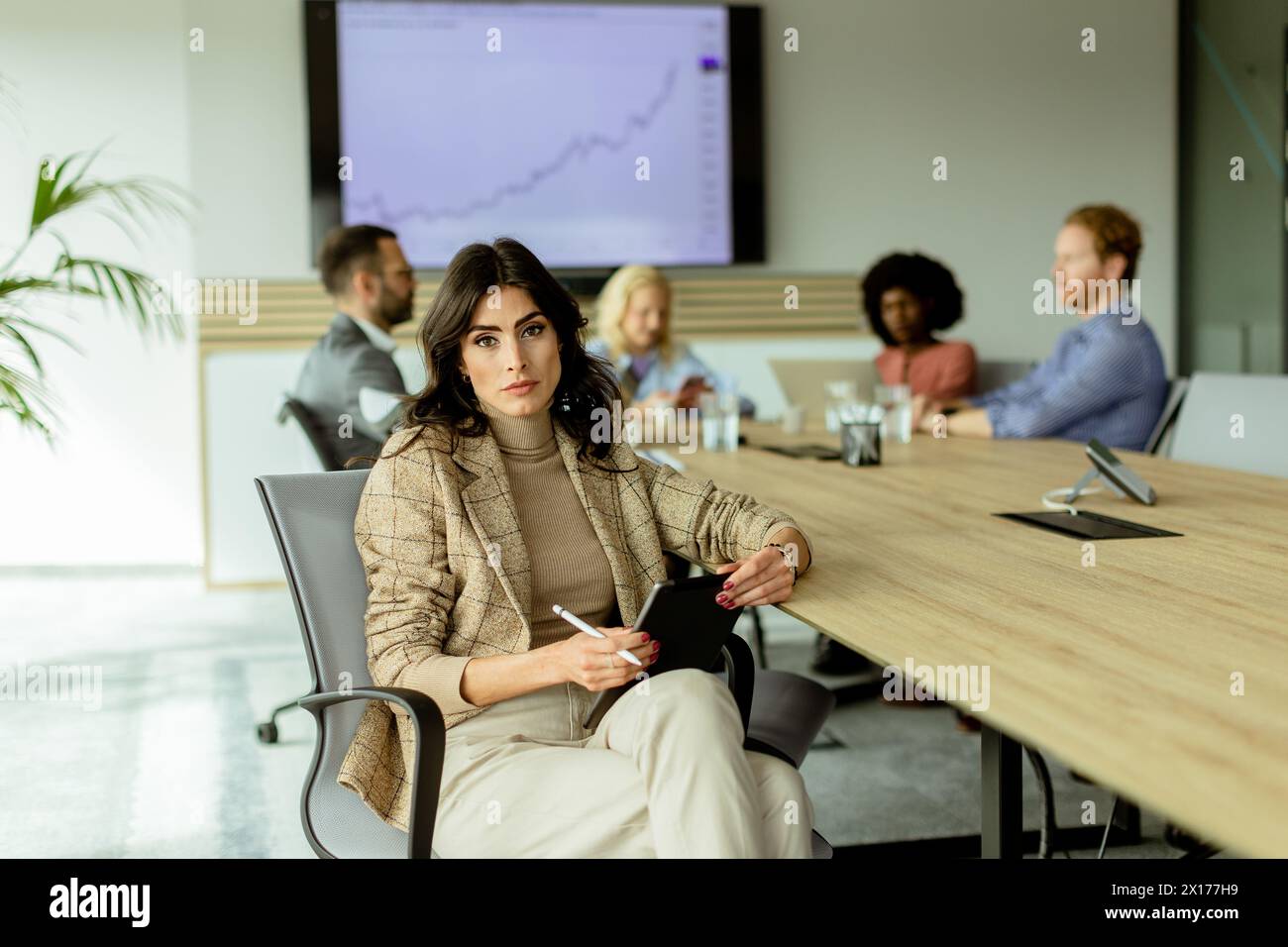 Femme d'affaires concentrée avec un bloc-notes réfléchit lors d'une réunion d'équipe, avec des collègues engagés dans la discussion en arrière-plan Banque D'Images