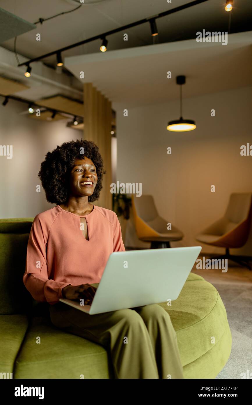 Femme d'affaires afro-américaine animée souriante tout en travaillant sur son ordinateur portable, confortablement assise dans un salon de bureau animé. Banque D'Images