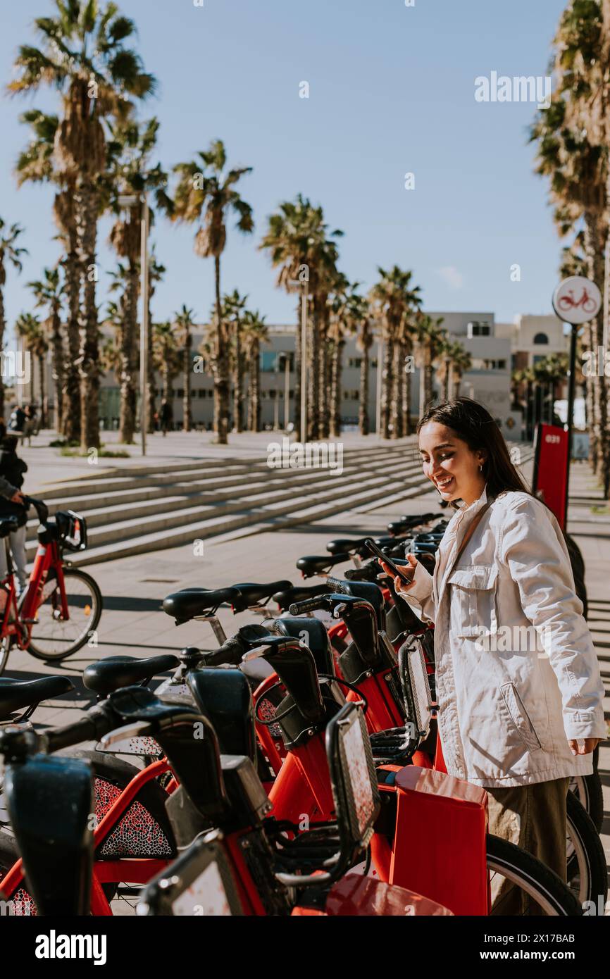 Jeune femme se tient près d'une rangée de vélos rouges dans la ville ensoleillée de Barcelone, vérifiant son téléphone, probablement en utilisant une application de partage de vélos Banque D'Images