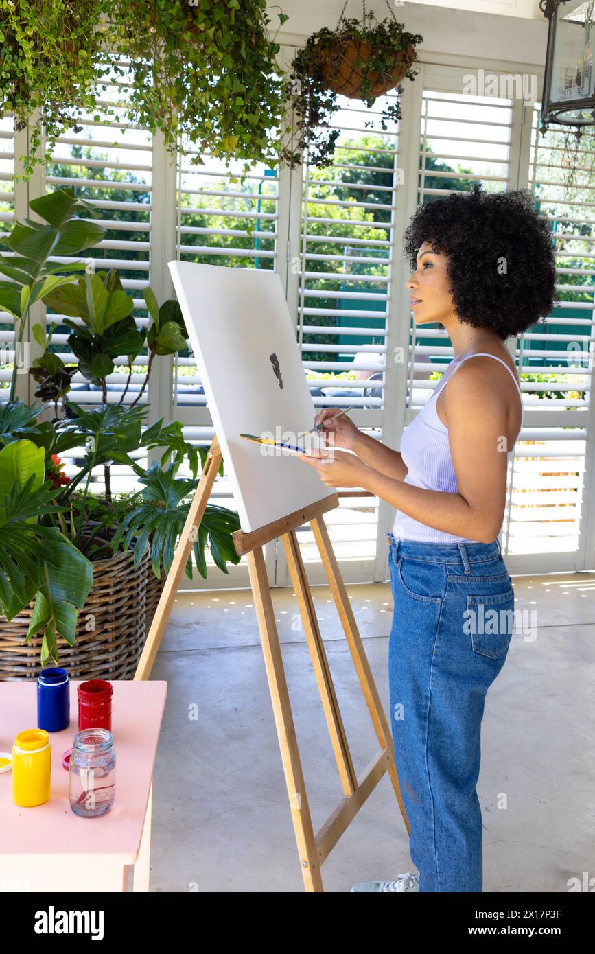 Jeune femme biraciale peignant sur toile à la maison, entourée de plantes Banque D'Images