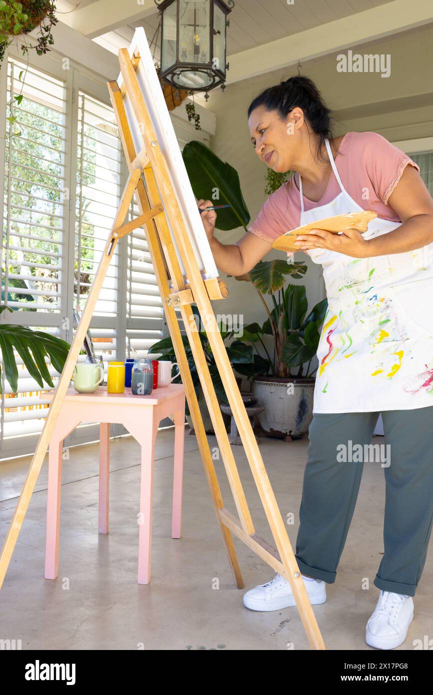 Femme biraciale mature peignant sur toile à la maison, portant un tablier Banque D'Images