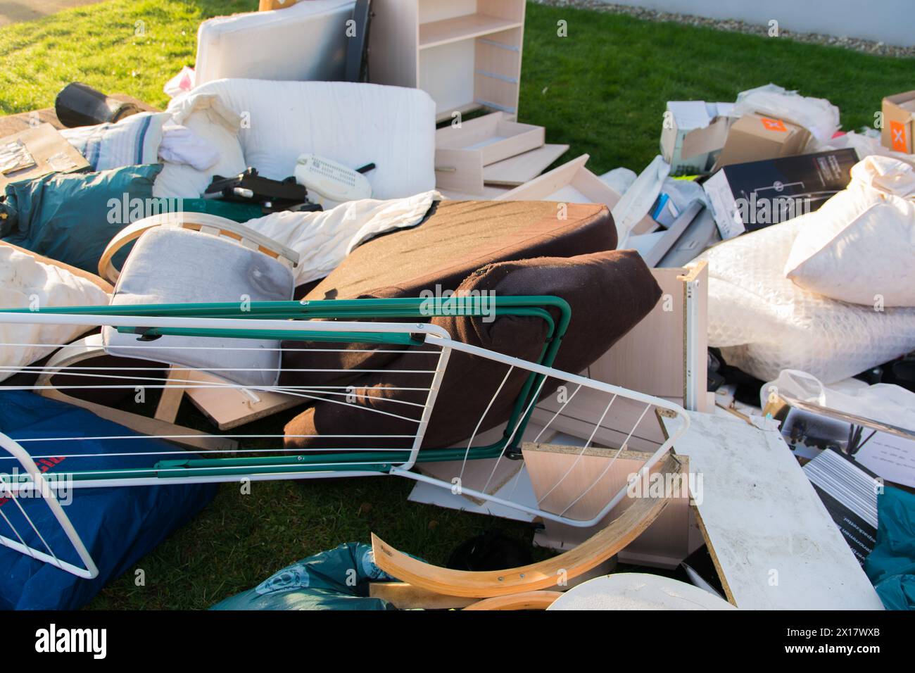 Beaucoup de déchets dans une ville près de la haus. Grandes ordures avec vieux meubles en bois, jouets, boîtes, sacs en plastique dans une rue et l'herbe verte au printemps ensoleillé tra Banque D'Images