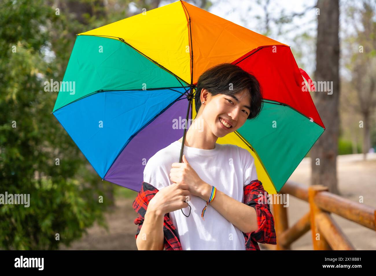 Mignon homme chinois gay souriant à la caméra tenant un parapluie couleur arc-en-ciel dans un parc Banque D'Images