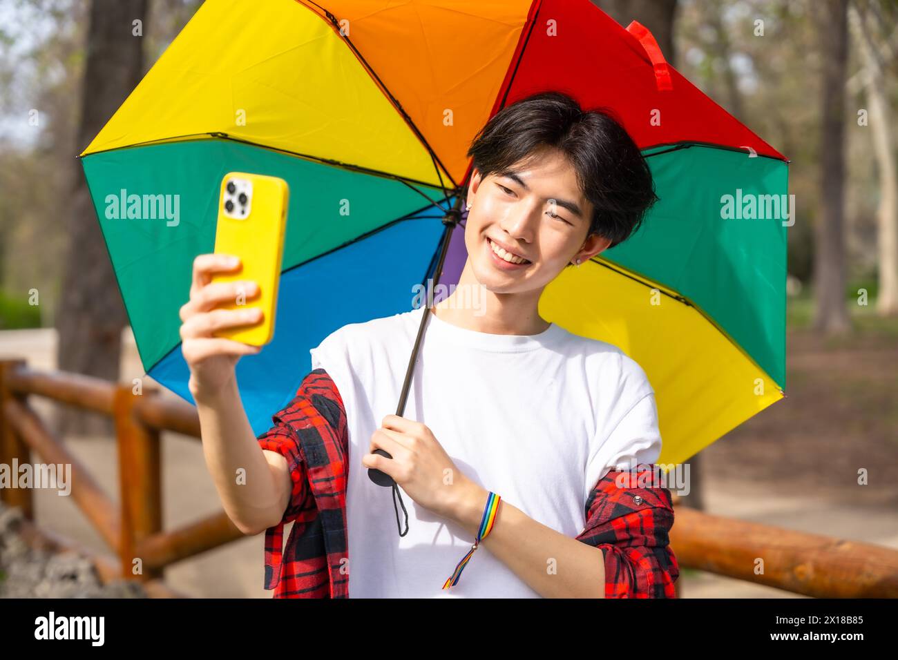 Homme gay chinois prenant un selfie avec un téléphone portable tout en tenant un parapluie de couleur arc-en-ciel debout dans un parc Banque D'Images