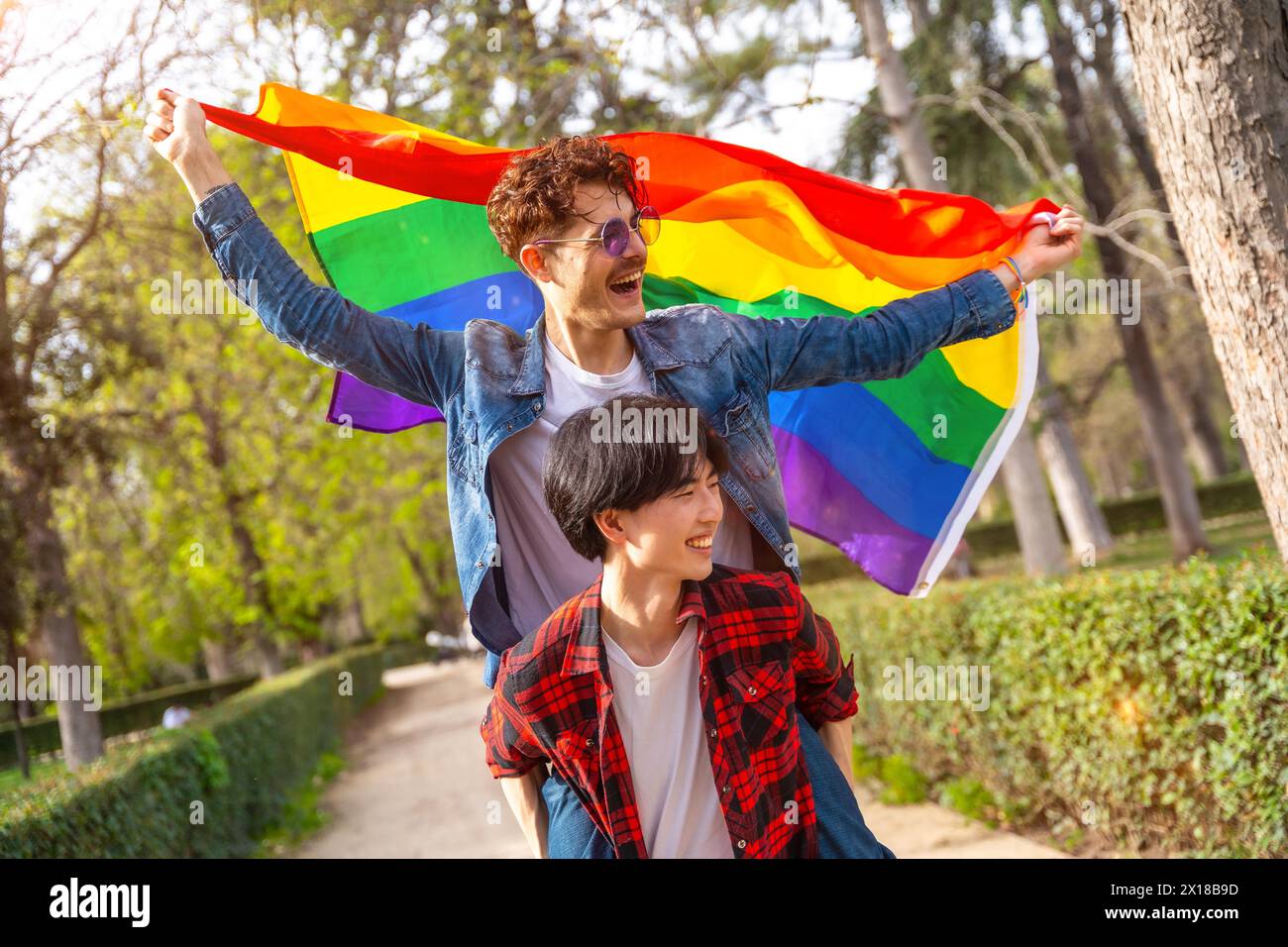 Couple gay multiracial agitant le drapeau lgbt piggyback célébrant la diversité dans un parc urbain Banque D'Images