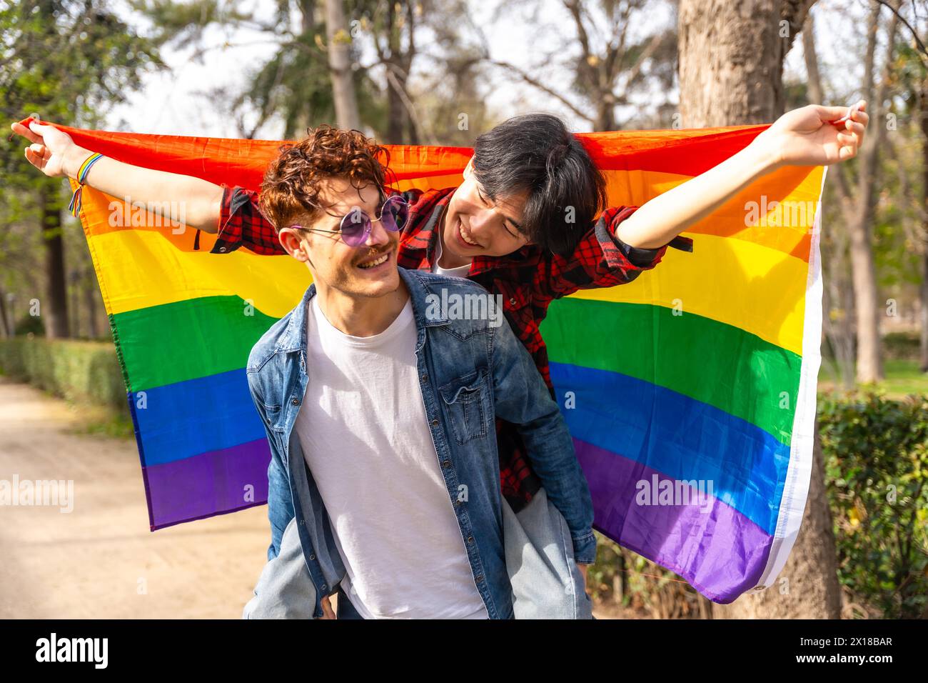 Portrait d'un joyeux couple gay multi-ethnique agitant le drapeau lgbt piggyback dans un parc Banque D'Images