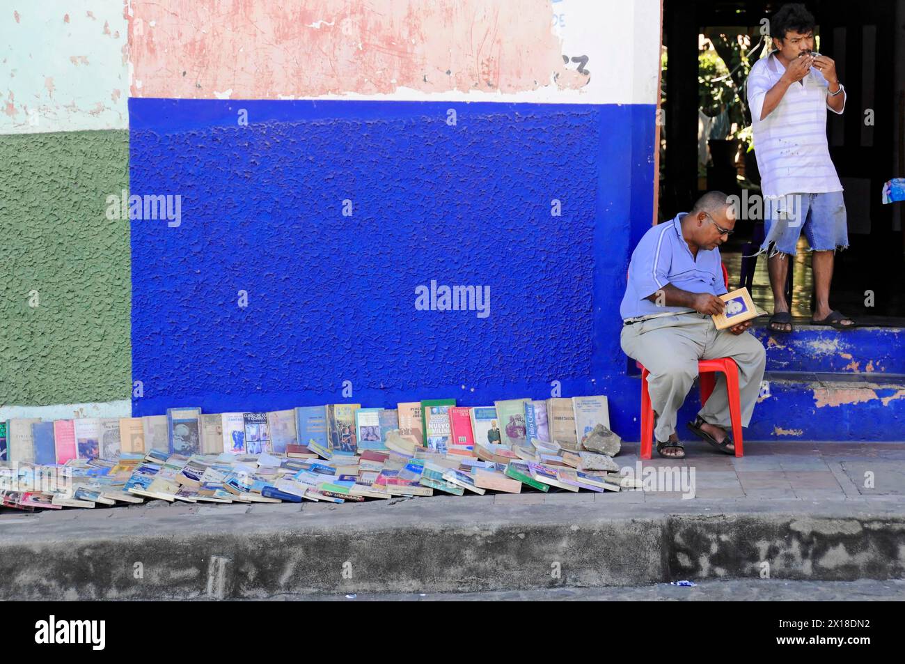 Leon, Nicaragua, Un vendeur de rue est assis devant un mur bleu vif vendant des livres usagés tandis que quelqu'un se tient à l'arrière-plan, Amérique centrale Banque D'Images