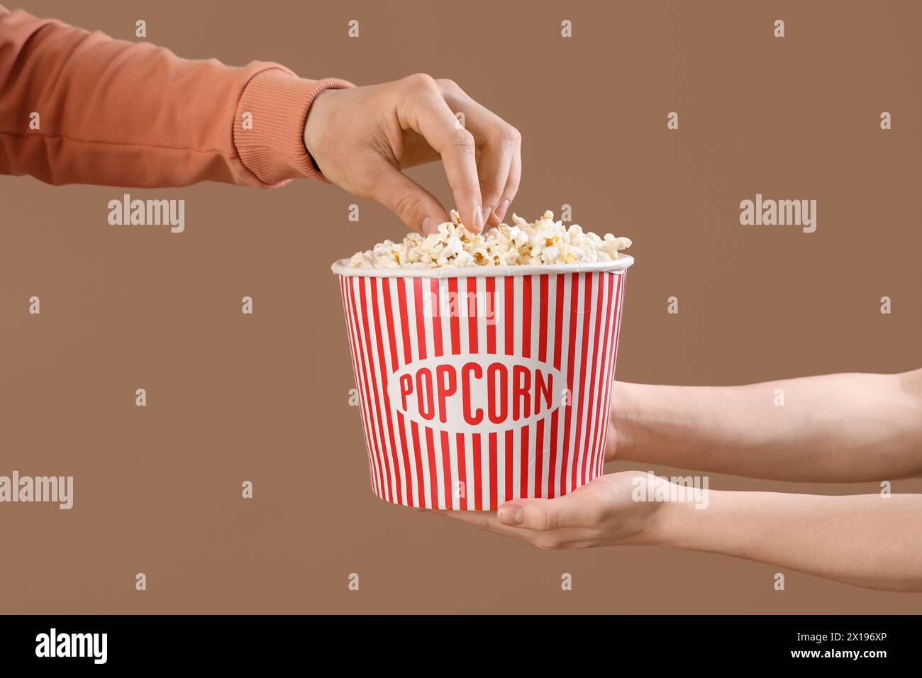 Main masculine prenant du pop-corn à partir de mains femelles de seau tenant sur fond brun Banque D'Images