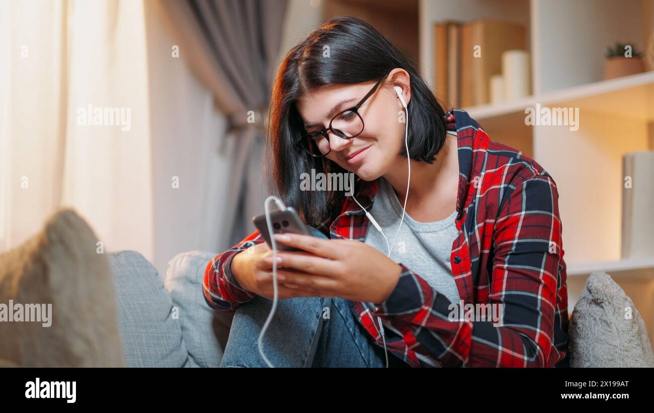 Conversation téléphonique. Loisirs gadgets. Femme millénaire heureuse tapant un message sur smartphone écoutant la chanson dans l'intérieur lumineux de la maison des écouteurs. Banque D'Images
