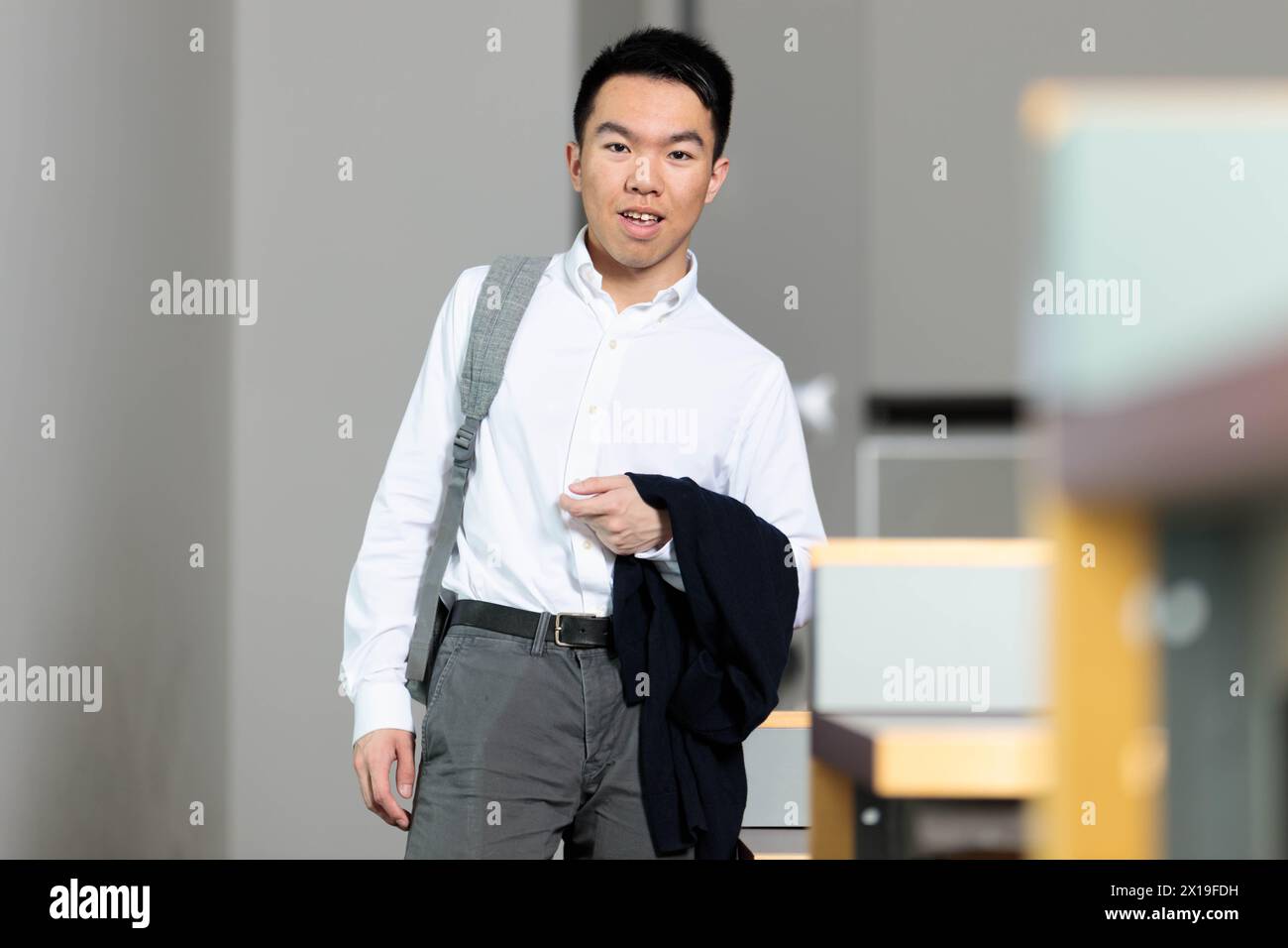 Un étudiant en affaires universitaire asiatique masculin dans une chemise habillée et un pantalon gris marche dans un couloir, il est prêt pour un événement formel dans la salle Banque D'Images