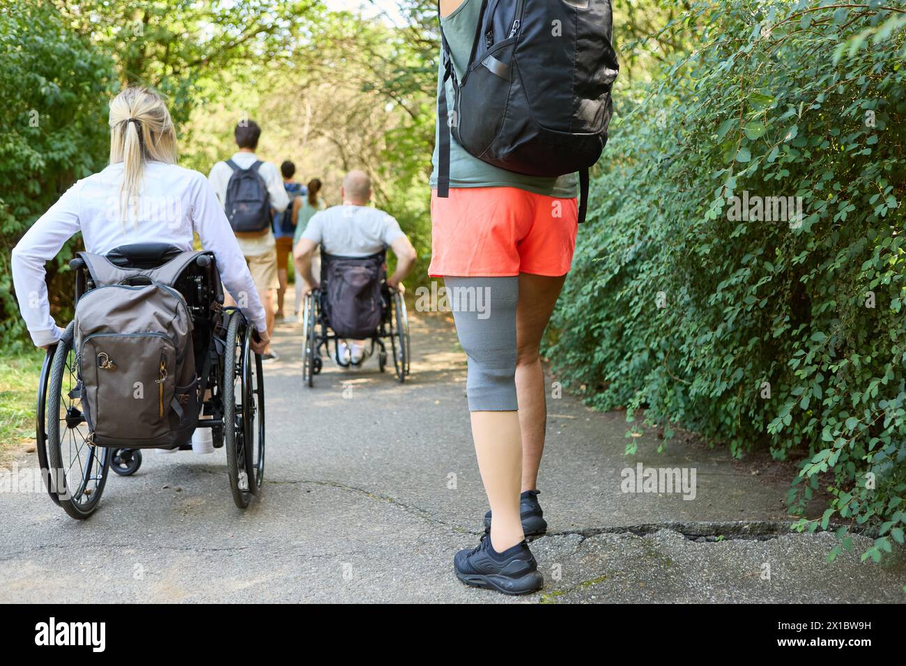 Une scène extérieure sereine capturant un groupe d'amis, deux en fauteuil roulant et une femme avec une prothèse de jambe, profitant d'une journée ensoleillée sur un sentier du parc Banque D'Images