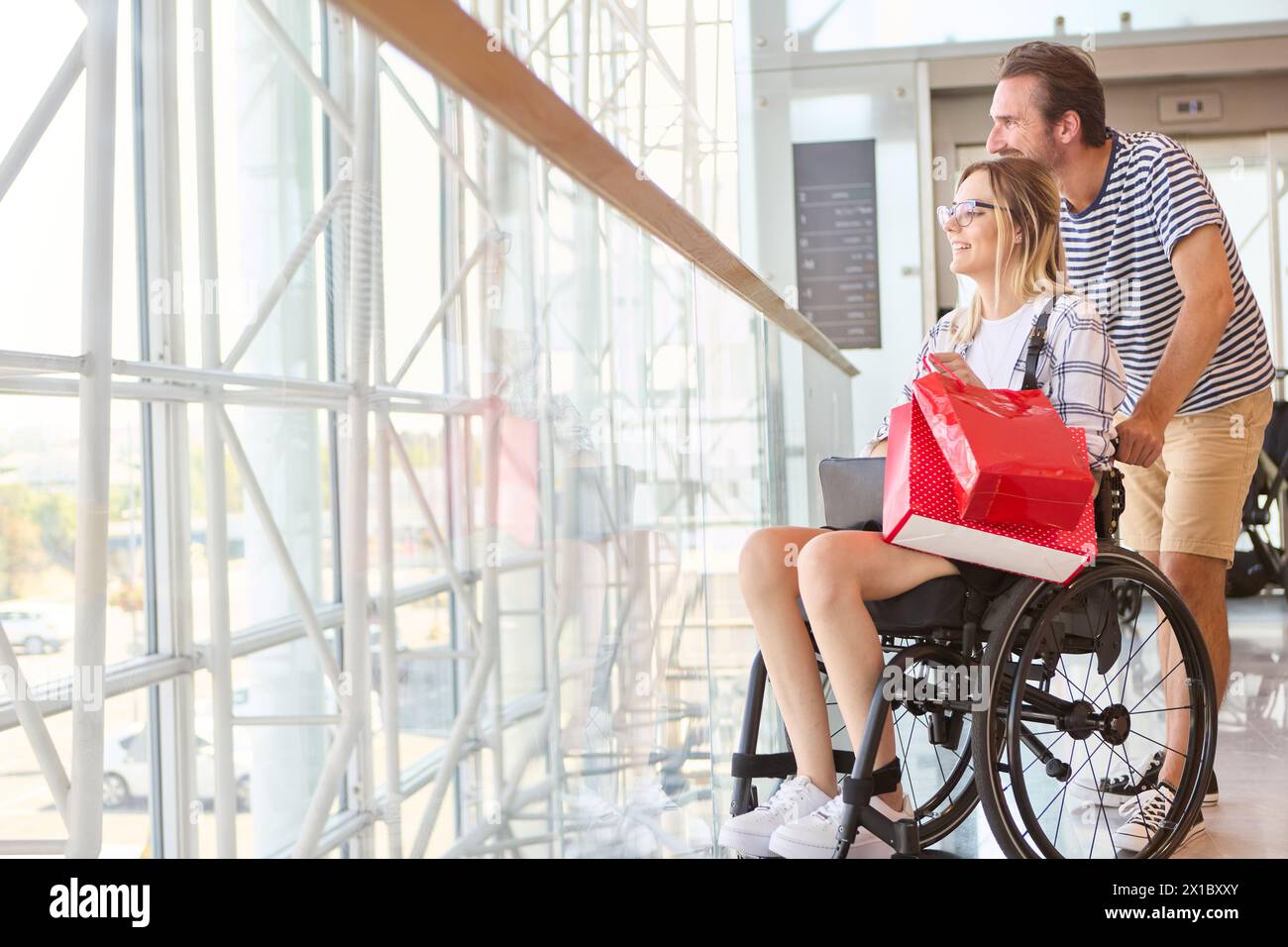 Un homme debout à côté d'une femme heureuse dans un fauteuil roulant, qui tient un cadeau rouge, dans un cadre ensoleillé et spacieux. Banque D'Images