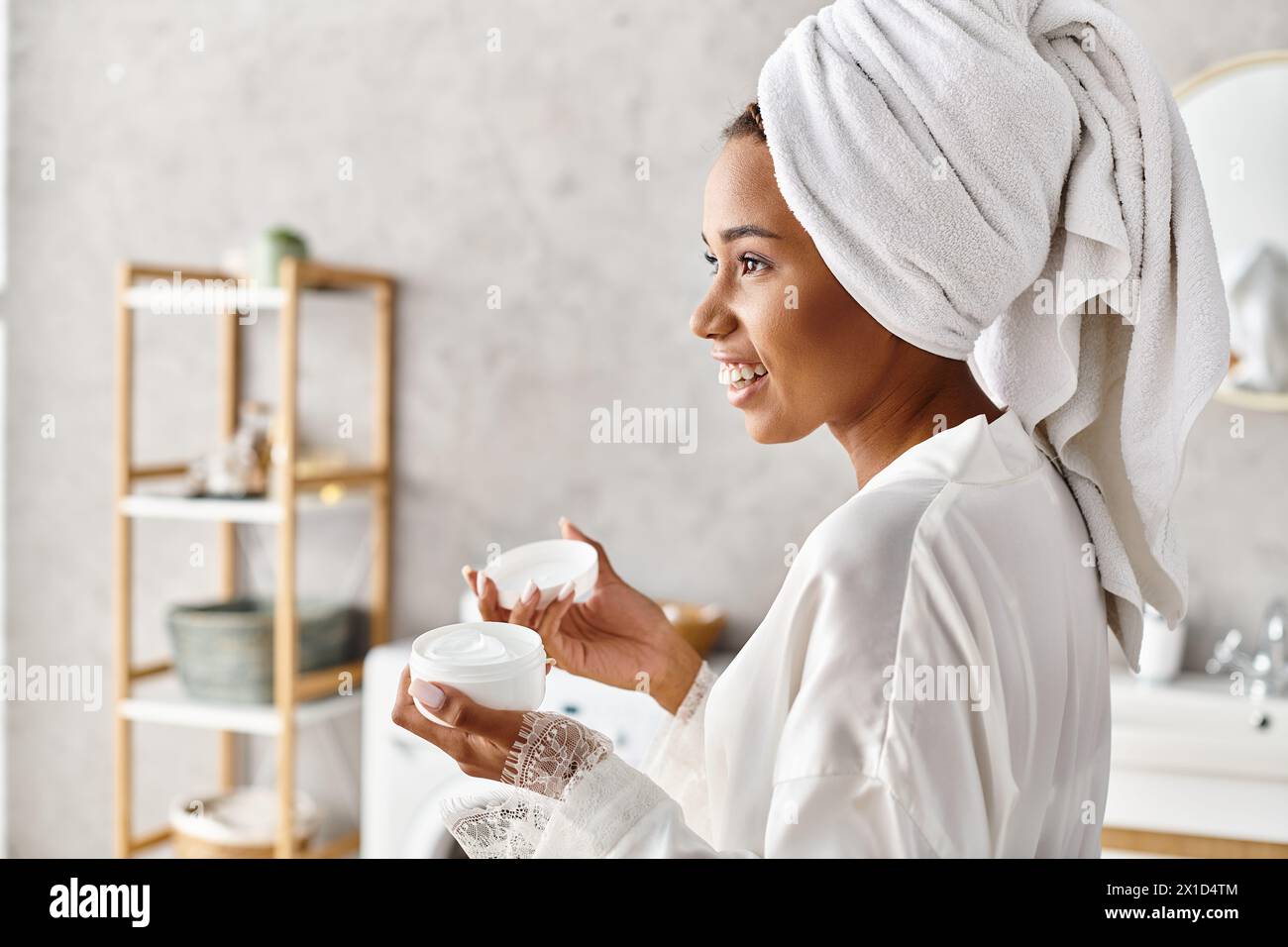 Une femme, avec une serviette sur la tête, tenant un pot avec de la crème dans sa salle de bain, profitant d'un moment de détente et de tranquillité. Banque D'Images