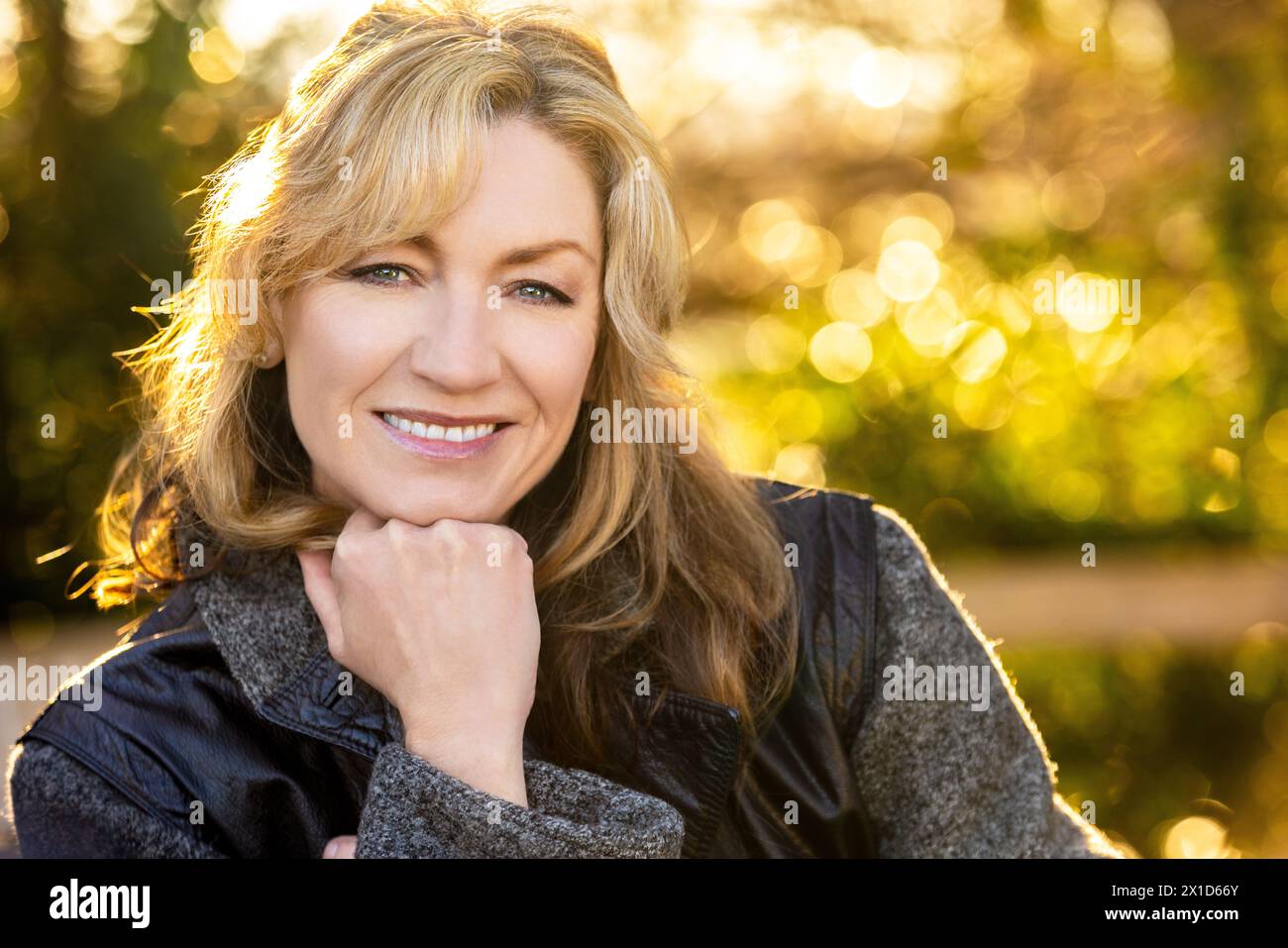 Belle femme d'âge moyen souriante heureuse à l'extérieur reposant sur sa main arrière éclairée par le soleil chaud au coucher ou au lever du soleil Banque D'Images